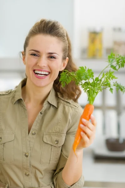 Happy young housewife holding carrot in kitchen — Stock Photo, Image