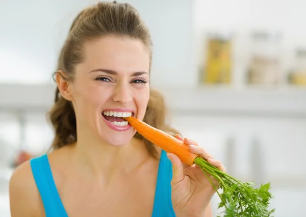 Happy young woman eating carrot in kitchen — Stock Photo, Image