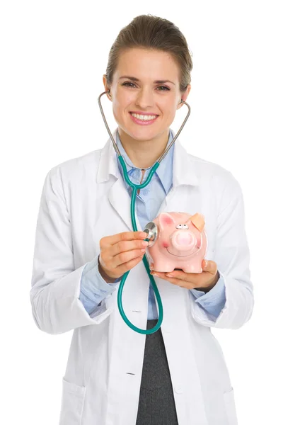 Smiling medical doctor woman listening piggy bank with stethosco — Stock Photo, Image