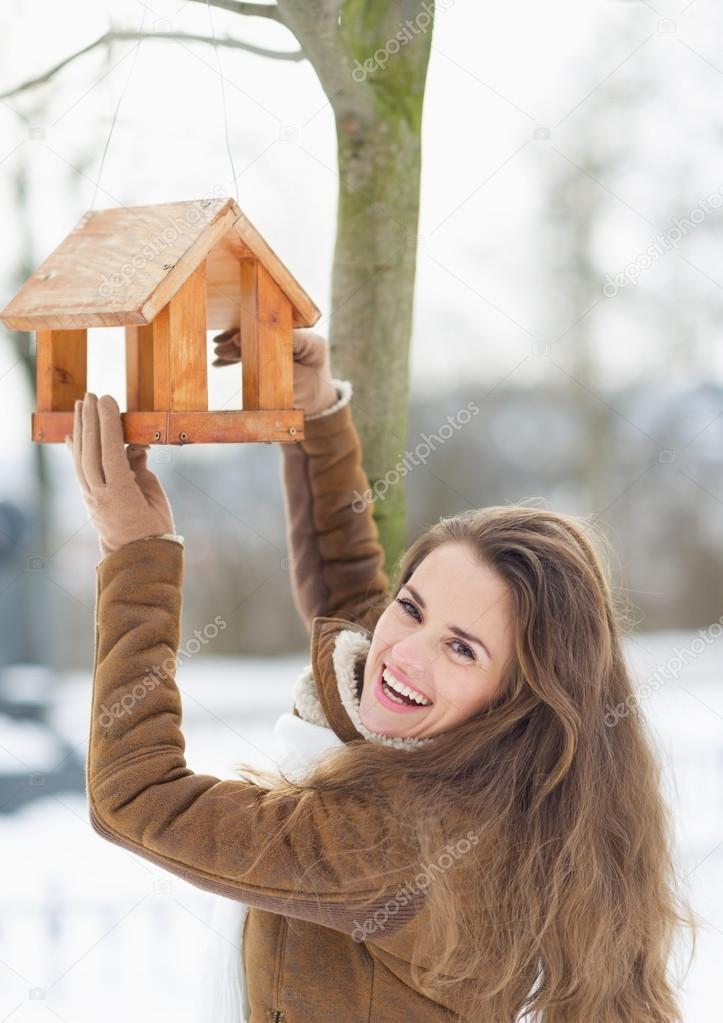Smiling young woman hanging bird feeder on tree in winter outdoo
