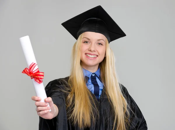 Smiling young woman in graduation gown showing diploma on grey b — Stock Photo, Image