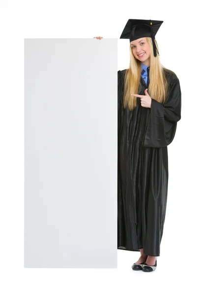 Mujer joven feliz en vestido de graduación apuntando en la cartelera en blanco — Foto de Stock