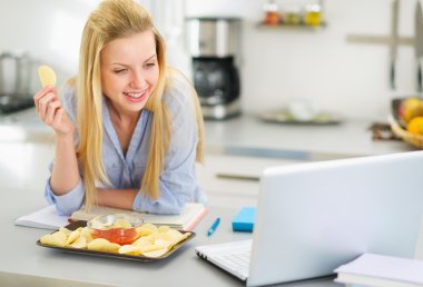 Teenage girl eating chips and looking in laptop clipart