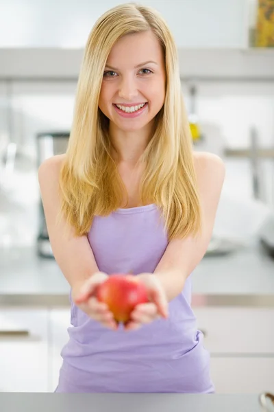 Smiling teenager girl giving apple in kitchen — Stock Photo, Image