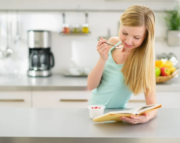 Счастливая девочка-подросток читает книги и ест йогурт на кухне — стоковое фото