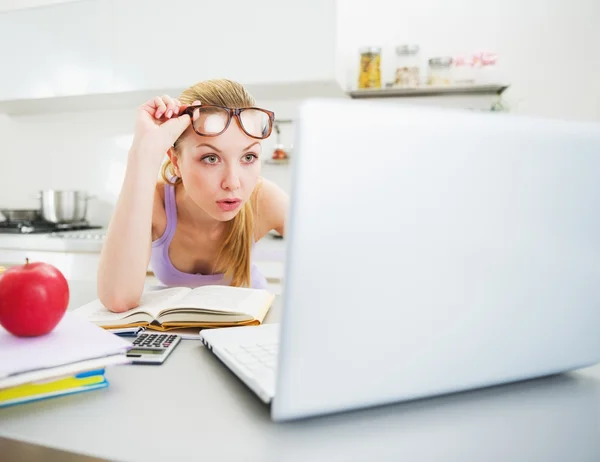 Überraschte junge Frau schaut in Laptop, während sie in Küche studiert — Stockfoto