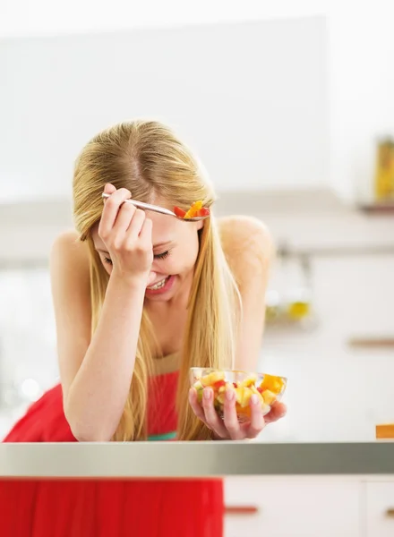 Lachen jonge vrouw vers fruit salade eten in keuken — Stockfoto
