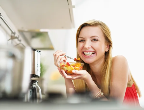 Mujer joven sonriente comiendo ensalada de frutas frescas en la cocina —  Fotos de Stock