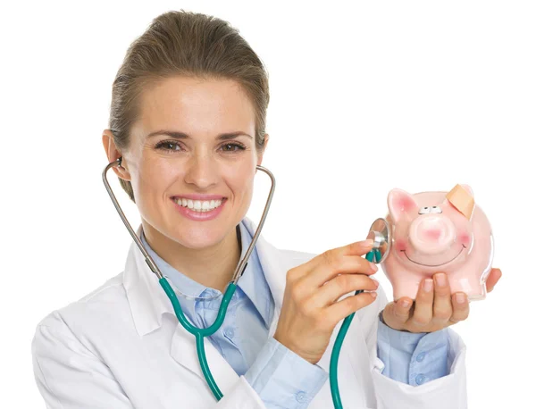 Lachende arts vrouw luisteren piggy bank met stethoscoop — Stockfoto