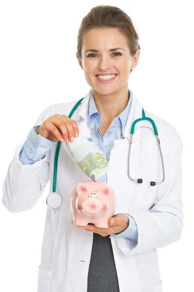 Femme médecin souriante mettant des billets en euros dans une tirelire — Photo