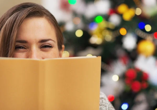 Glückliche junge Frau versteckt sich hinter Buch neben Weihnachtsbaum — Stockfoto