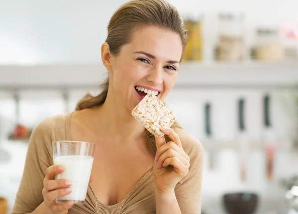 Happy young woman eating crisp bread with milk in kitchen — Stock Photo, Image