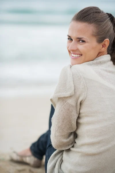 Улыбающаяся молодая женщина в свитере сидит на одиноком пляже — стоковое фото