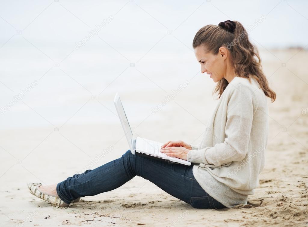 Woman in sweater sitting on beach with laptop