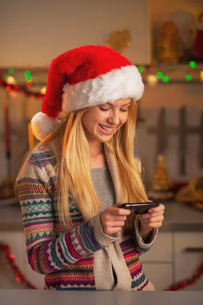 Smiling teenage girl in santa hat writing sms in christmas decorated kitchen — Stock Photo, Image