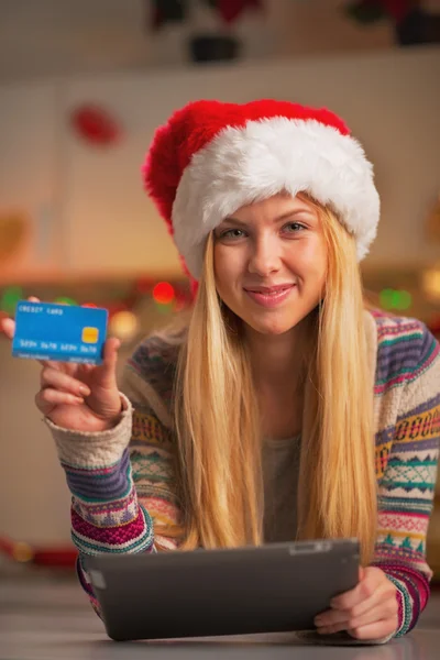 Smiling teenage girl in santa hat with tablet pc showing credit card — Stock Photo, Image