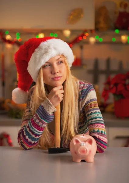 Portrait of thoughtful teenage girl in santa hat with hammer and piggy bank — Stock Photo, Image