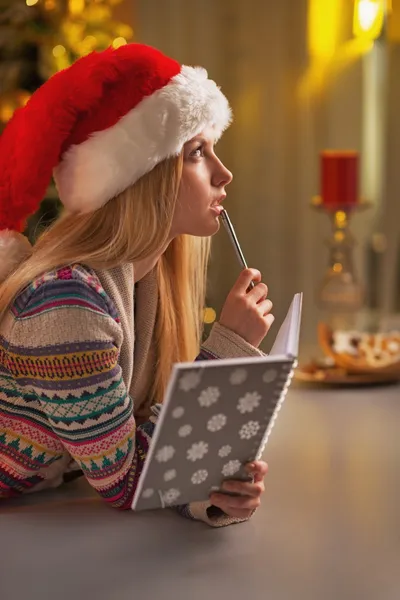 Ragazza adolescente pensierosa in cappello di Babbo Natale con diario in cucina decorata di Natale — Foto Stock