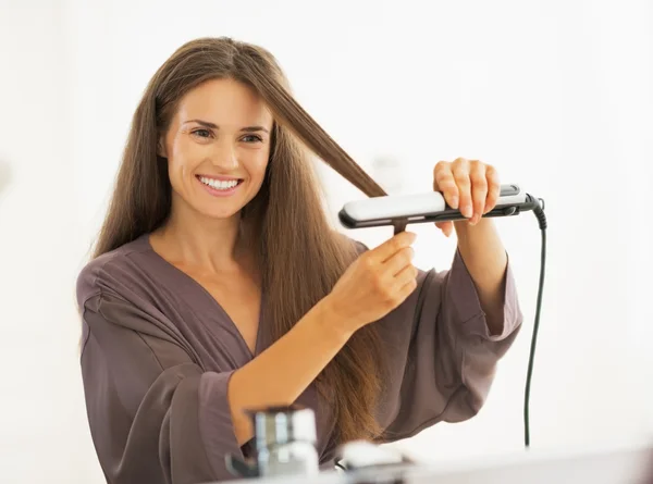 Happy young woman straightening hair with straightener — Stock Photo, Image