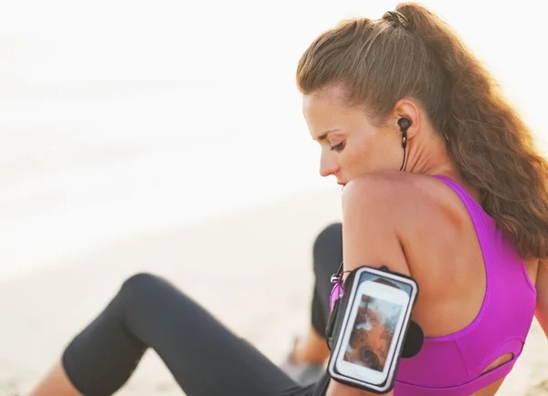 Thoughtful fitness young woman in headphones sitting on beach — Stock Photo, Image