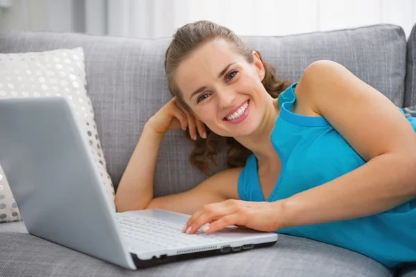 Mujer joven sonriente acostada en un sofá con computadora portátil —  Fotos de Stock