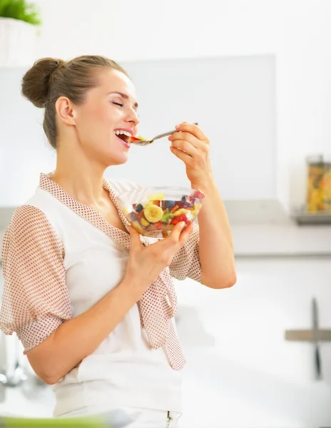 Gelukkig jonge huisvrouw eten van verse fruitsalade in keuken — Stockfoto