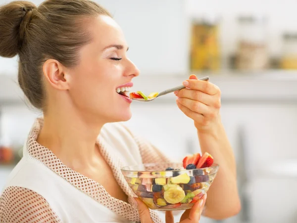 Jovem dona de casa feliz comendo salada de frutas frescas na cozinha — Fotografia de Stock