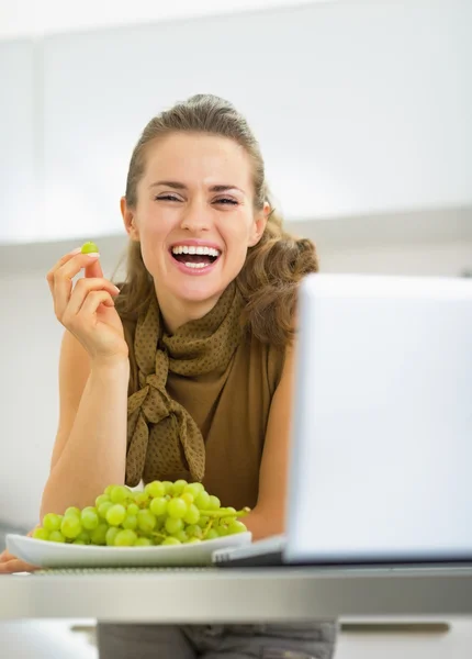 Jovem dona de casa feliz com laptop comendo uva na cozinha — Fotografia de Stock
