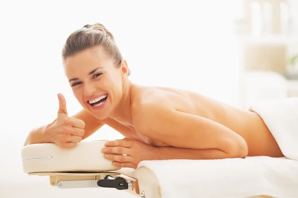 Smiling young woman laying on massage table and showing thumbs up — Stock Photo, Image