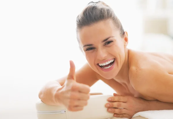 Lächelnde junge Frau liegt auf Massagetisch und zeigt Daumen hoch — Stockfoto