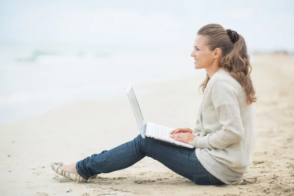 Ευτυχισμένη γυναίκα που κάθεται με το laptop στο κρύο παραλία — Φωτογραφία Αρχείου