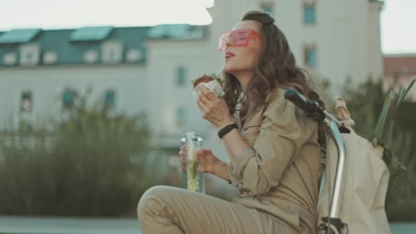 サングラスをかけ街の外に座っている間に文字列バッグ スクーター サンドイッチを食べながら全体的にエレガントな中年の女性 — ストック動画