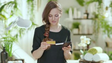 Gülümseyen 40 yaşındaki kadın çalışan akıllı telefon ve kredi kartı ile modern güzellik salonunun e-ticaret sitesinde online alışverişler yapıyor..