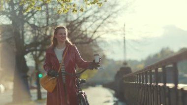 Kırmızı yağmurluklu, bisikletli ve kahveli zarif bir kadın şehrin dışında yürüyor..