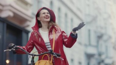 Gülümseyen 40 yaşındaki kırmızı yağmurluk giymiş, yağmurda dışarıda bisiklet süren bir kadın..