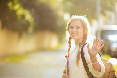 smiling stylish girl in white sweatshirt with backpack hand waving and going to school outdoors in the city.