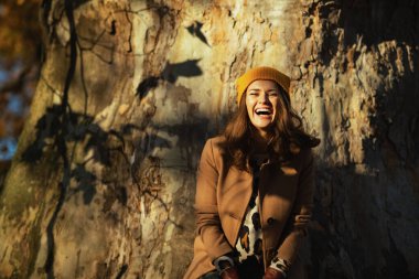 Merhaba Ekim. Kahverengi ceketli, sarı şapkalı, gülen modern kadın sonbaharda şehir parkında büyük bir ağaca karşı..