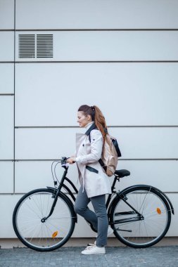 Şehrin dışındaki metal bir duvarın önünde bej renkli trençkotlu ve sırt çantalı gülümseyen zarif kadının tam boy portresi..