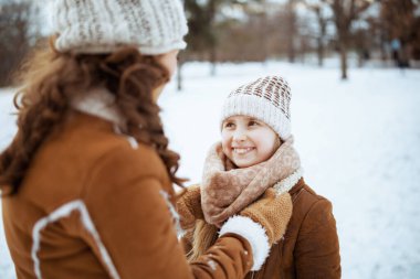 happy modern mother and daughter in a knitted hats and sheepskin coats with mittens in a knitted hat and sheepskin coat outdoors in the city park in winter.