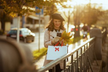 Hello november. happy modern middle aged woman in beige trench coat with shopping bags and autumn yellow leaves using smartphone applications outdoors on the city street in autumn.