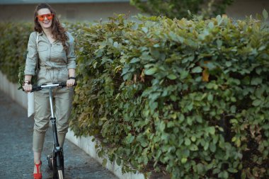 Mutlu, orta yaşlı, güneş gözlüklü, spor çantalı ve motosikletli bir kadın şehrin yeşil çitlerinin yanında..