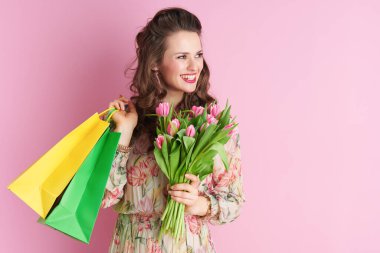 happy middle aged woman in floral dress with tulips bouquet and shopping bags isolated on pink background.