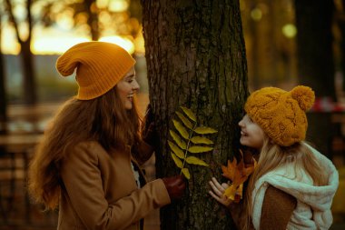 Merhaba Ekim. Sonbaharda şehir parkında sonbahar sarısı yapraklarıyla turuncu şapkalı gülümseyen anne ve kız..