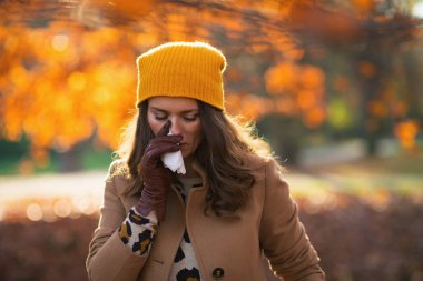 Hello october. tired trendy middle aged woman in brown coat and yellow hat with napkin blowing nose outdoors on the city park in autumn.