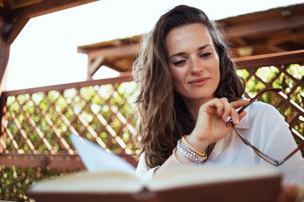 Pensive Modern Woman White Shirt Eyeglasses Reading Book Patio Guest — Stok Foto