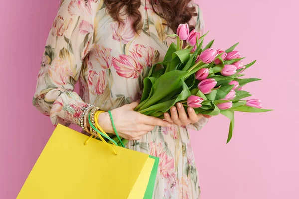 Closeup on middle aged woman with tulips bouquet and shopping bags isolated on pink background.