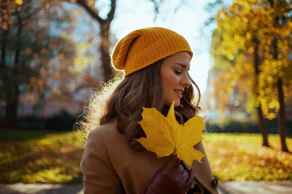 Hello november. elegant 40 years old woman in beige coat and orange hat with autumn yellow leaves outside in the city in autumn.