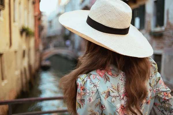 Seen from behind modern woman in floral dress with hat exploring attractions in Venice, Italy.