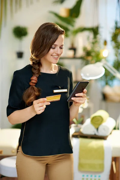 Uśmiechnięta Letnia Pracownica Smartfonem Kartą Kredytową Kupująca Online Nowoczesnym Salonie — Zdjęcie stockowe