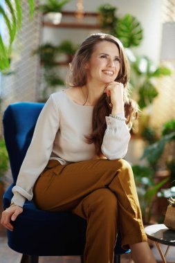 Green Home. happy trendy middle aged housewife with long wavy hair in the modern house in sunny day in green pants and grey blouse sitting in a blue armchair.